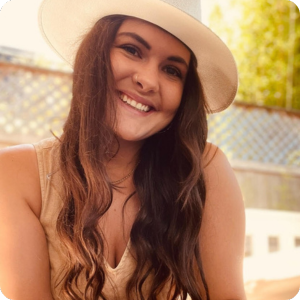 Untitled design (14) A woman wearing a sleeveless dress and hat is smiling at the camera with a house and tree leaves in the background.