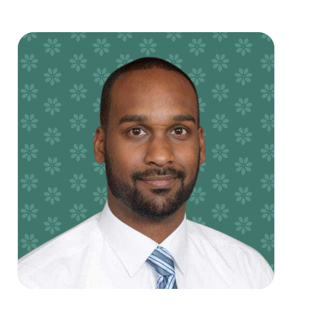 ian Portrait of a smiling therapist at Total Life with a neatly trimmed beard and short black hair, wearing a white dress shirt and a blue striped tie. The background features a muted teal color with a subtle floral pattern.