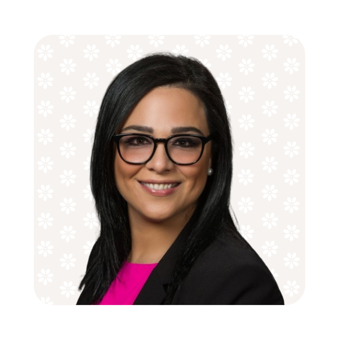 tanya Portrait of a smiling therapist at Total Life with light skin and straight black hair, wearing black-framed glasses, a black blazer, and a vibrant pink blouse. The background features a soft beige color with a subtle floral pattern.
